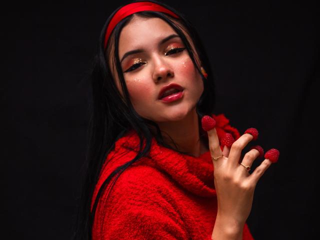 LizzieJoy a de longs cheveux noirs, un serre-tête rouge et un sweat à capuche rouge, avec des yeux rouges pailletés et du rouge à lèvres rouge. Elle touche son visage avec ses ongles peints en rouge, dégageant une tension sensuelle. Fond noir.