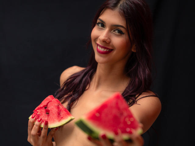 DanikaSerov, avec de longs cheveux noirs, souriante, tenant deux tranches de pastèque juteuses, une dans chaque main, sur fond noir. fond, sans vêtements, rouge à lèvres rouge vif, séduisant, tension sexuelle.