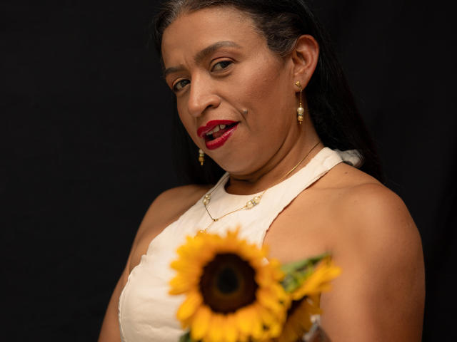 Fotografia di SoffieMature con pelle marrone media, capelli scuri raccolti, che indossa una canotta bianca, orecchini dorati. rossetto rosso, che tiene un girasole, sorridente, sfondo scuro, sguardo intenso, sexy.