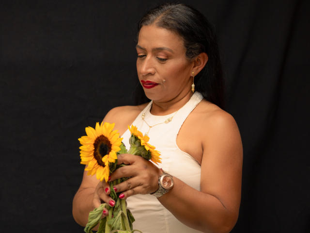 SoffieMature, avec sa peau bronzée et son rouge à lèvres rouge, tient deux tournesols, et porte un haut blanc à encolure dégagée ainsi qu'une montre en or. Elle regarde en bas avec un léger sourire, ses cheveux noirs lissés en arrière..