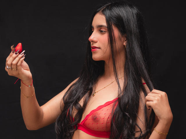 TiffannyRose, com longos cabelos pretos, vestindo um sutiã de renda vermelho, segurando uma morango vermelha perto do rosto. Ela tem pulseiras de ouro e um anel. Fundo preto. Incrivelmente sexy..