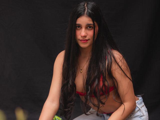 TiffannyRose, con lunghi capelli neri e bagnati, si sporge in avanti, indossando un reggiseno rosso e pantaloni bianchi, sfondo scuro, sguardo intenso. un leggero sorriso, scollatura visibile, gocce d'acqua nei capelli, posa seducente.