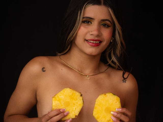 SheylaSpooner, sem blusa e com pele clara, segurando dois objetos amarelos, fofos e redondos em frente aos seus mamilos. Ela tem cabelo preto, batom vermelho, uma tatuagem de uma lua crescente no ombro esquerdo e um colar de ouro..