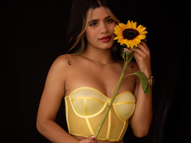Sheyla Spooner, com longos cabelos escuros, segurando um girassol no rosto, veste um corpete amarelo sem alças e tem uma pequena tatuagem. Em seu ombro direito, contra um fundo preto. Muito sexy..