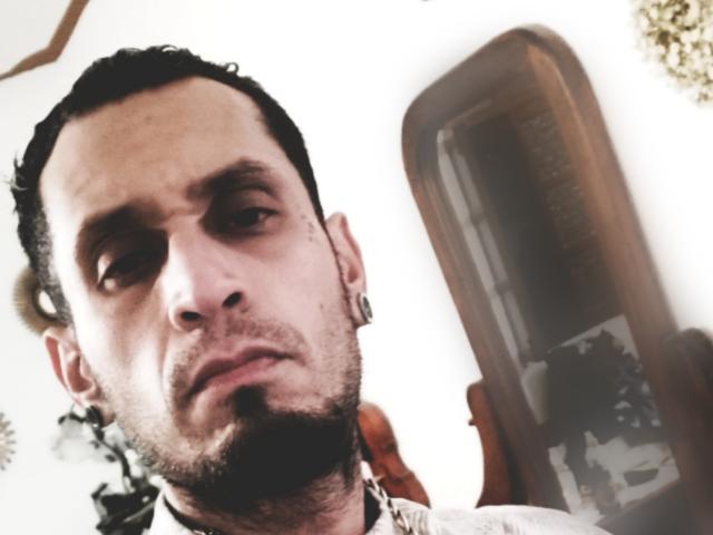 Close-up foto van ZousGreen, een man met een baard, kort zwart haar, een intense blik en een schaduwrijk gezicht. Hij draagt een wit shirt. Aan zijn rechterkant staat een camera, die licht weerkaatst..