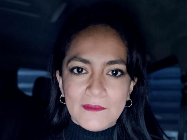 Close-up photograph of AnttonellaX69, a Latina woman with dark hair, red lipstick, and hoop earrings. She has intense black eyes and a serious expression. Dim lighting creates shadows on her face.