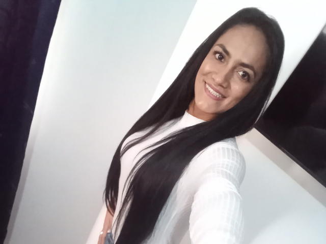 Photograph of AnttonellaX69 with long black hair, wearing a white shirt, smiling at the camera. Bright white background, dark edges. Sexy, attractive, light brown skin, dark eyes, subtle makeup.