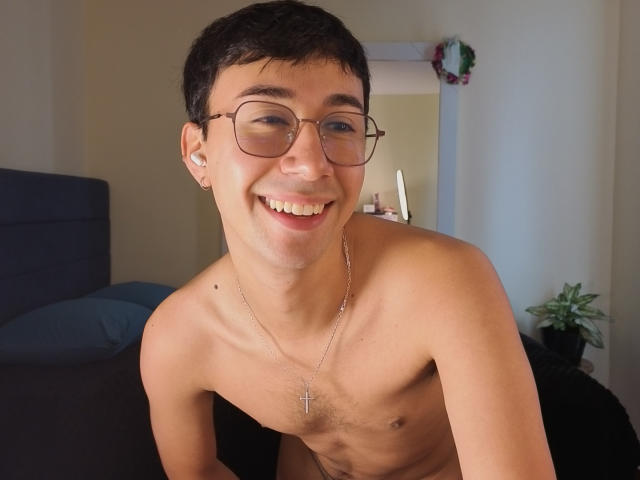 Photograph of a shirtless young man with short black hair, glasses, and a cross necklace, leaning forward, smiling, showing his teeth, with visible arousal, flaccid cock hinting at sexual tension.