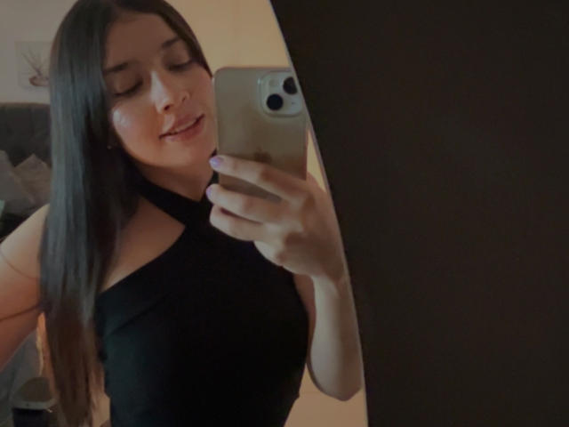 Photograph of EmmaLombardi, taking a selfie with a smartphone. She has long black hair, a black halter top, and is smiling slightly. Her left hand holds the phone, covering part of her chest.