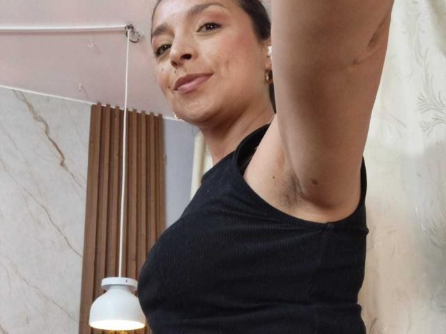 Close-up photo of StangelarRivera with dark skin, black tank top, raised arm, armpit visible, slight smirk, indoor setting, marble wall, brown curtains, hanging lamp, subtle sexual tension.