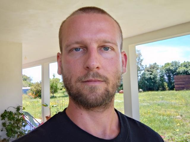 Close-up photo of ExhibBoby, a bald, bearded man with blue eyes, wearing a black shirt. Bright outdoor setting with green grass, trees, and white columns in the background. Fucking serious expression.