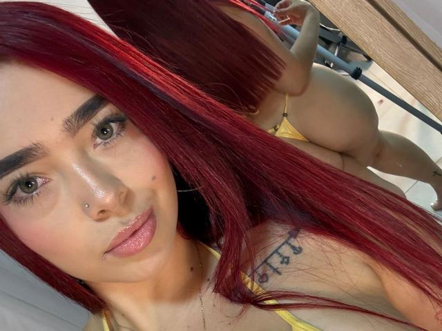 Close-up photo of ValeriaRoseT with red hair, green eyes, and a nose piercing. She's wearing a yellow bikini, her ass is raised and prominent. Her tattoo is visible on her shoulder. Her skin is shiny.