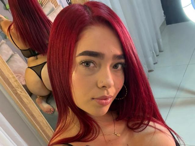 ValeriaRoseT, con lunghi capelli rossi, indossa uno slip nero, e il suo riflesso è visibile nello specchio. Il viso di ValeriaRoseT in primo piano, i glutei riflessi nello specchio. Ha un piercing al naso, orecchini a cerchio e una collana. Molto attraente..