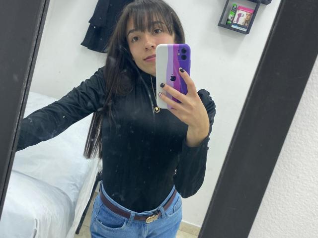 Selfie of ElodiePerrine, long black hair, bangs, wearing black turtleneck, blue jeans, holding purple phone, white background, black mirror frame, reflection, casual pose, sexy, slim physique, slight cleavage.