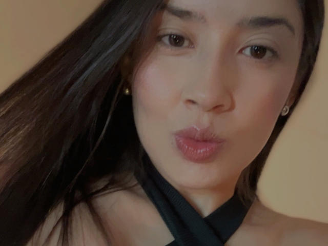 Nahaufnahme von Emma Lombardi, einer asiatischen Frau mit langen, schwarzen Haaren, voller Lippen und braunen Augen, die ein schwarzes Choker-Halsband trägt. Ihre Haut ist glatt, und sie macht ein Küssengesicht. Verdammt sexy..
