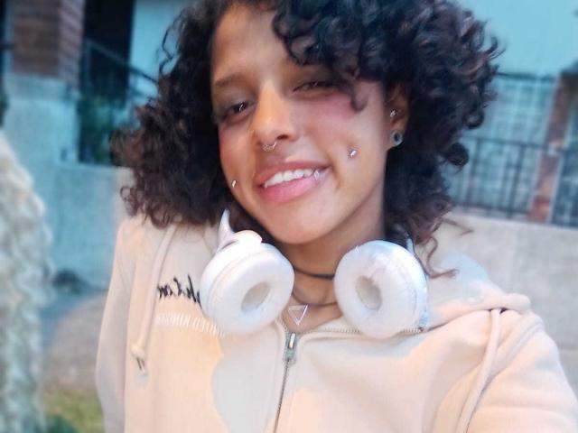 Close-up of LunaKeller, a curvy woman with dark curly hair, smiling, wearing a white hoodie, white headphones around her neck, nose ring, and earrings. Bright natural light, slight blue tint.