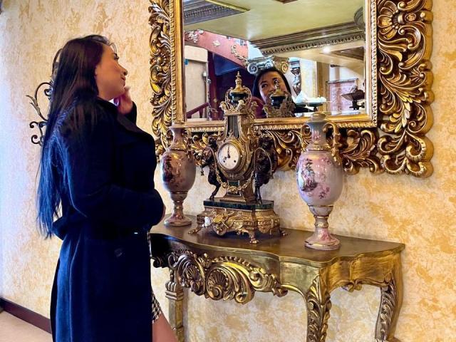 Karimelee, com longos cabelos pretos, vestida com um casaco preto, está ao lado de uma mesa de console dourada ornamentada com dois detalhes decorativos. Vasos. O reflexo em um espelho grande e ricamente emoldurado mostra seu rosto..