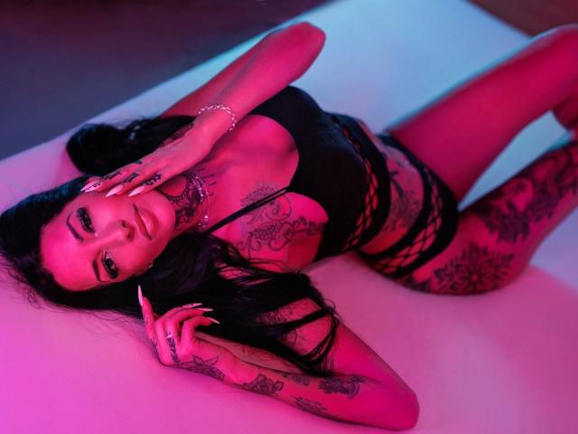 RubyValentine-hot, laying on a white surface, wearing black lace lingerie, heavily tattooed, pink and blue lighting, seductive pose, hand on face, dark hair, tattoos on arms and chest, sexy vibe.