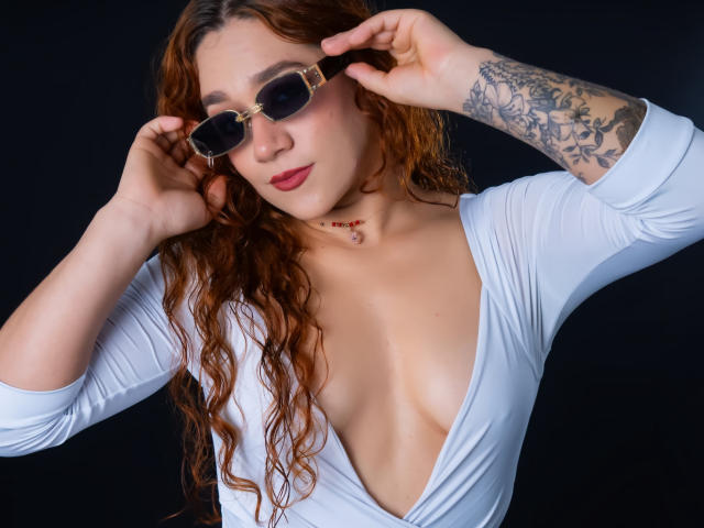 Valery Hilton, una donna formosa con lunghi capelli castani ondulati, indossa una camicetta bianca a scollo profondo che mette in risalto il decolleté, e lo sistema. Occhiali da sole neri.
braccio tatuato, rossetto rosso, sfondo scuro, sguardo intenso. Incredibilmente sexy..