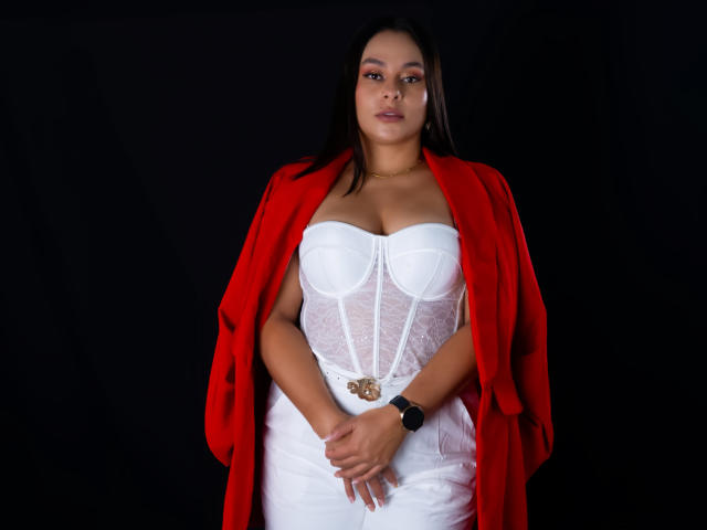 LunaSoler está de pie, centrada sobre un fondo negro, vistiendo un corpiño blanco de encaje y un abrigo rojo que lleva puesto sobre ella. hombros. Ella tiene una pulsera negra en su muñeca derecha, mirando directamente a la cámara con una expresión seria. Sus manos están entrelazadas frente a ella. Tiene el cabello oscuro y un collar de oro..