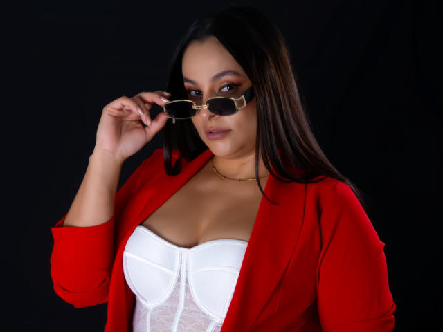 LunaSoler, with long black hair, wears a red cardigan over a white, strapless top. She holds black sunglasses to her face, exuding confidence and subtle sexual tension. Black background.