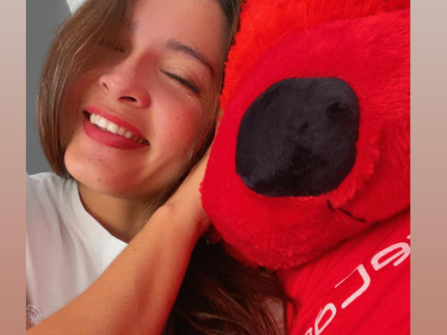 Close-up photo of Gabrielafoxxy smiling with eyes closed, hugging a bright red stuffed bear with a black nose and white text on the side. Her dark brown hair is loose. Fucking cute.