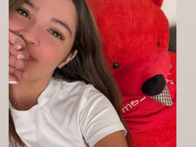 Gabrielafoxxy ligt naast een enorme rode teddybeer, haar gezicht gedeeltelijk verborgen door haar hand, met een suggestieve glimlach en donkere ogen. Ze draagt een witte blouse, met loshangend bruin haar..