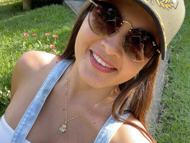 Gros plan de Gabrielafoxxy souriant, portant de grandes lunettes de soleil foncées, un haut blanc à encolure dégagée et un collier en or avec un cœur. pendant.
herbe baignée de soleil et fleurs rouges en arrière-plan. Légères marques de bronzage visibles..