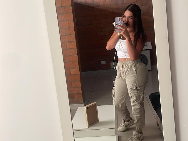 Gabrielafoxxy, una donna con i capelli castani, che si scatta un selfie allo specchio, indossa una canotta bianca, pantaloni cargo beige e stivali bianchi, e tiene in mano un... telefono viola, sfondo con muro di mattoni, scatola di cartone sul pavimento, posa casuale, sexy..