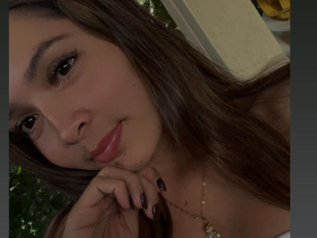 Nahaufnahme-Selfie von Gabrielafoxxy, einer braunhaarigen Frau mit dunklen Augen, roten Lippen, schwarzem Nagellack und einer goldenen Halskette. mit einem kleinen Anhänger. Sie legt ihr Kinn in ihre Hand und strahlt eine verführerische Ausstrahlung aus..