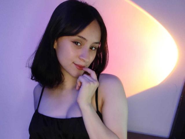 Fotografía de AnnieLamberth con cabello oscuro hasta los hombros, vistiendo una blusa negra con tirantes finos, tocándose ligeramente. Con una leve sonrisa en el rostro. El fondo presenta un arco de luz brillante y colorido. Increíblemente atractivo..