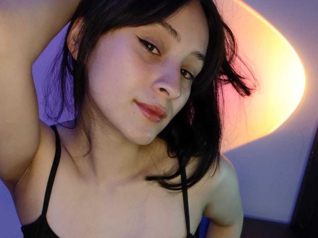Close-up of AnnieLamberth, an Asian woman with black hair, wearing a black spaghetti strap top, looking directly at the camera with a slight smirk, arm raised. Bright orange light in the background.