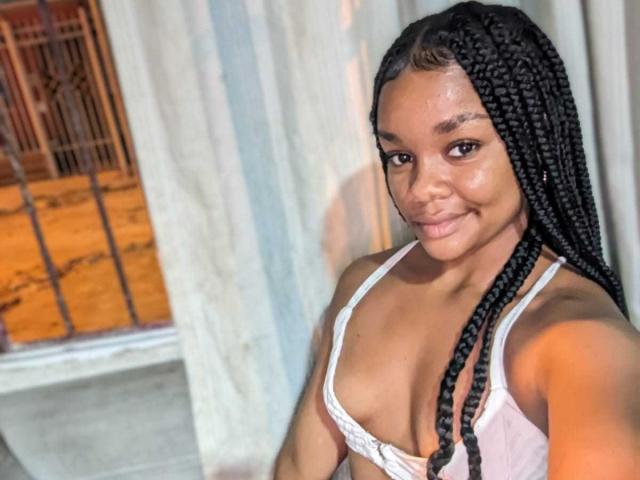 Fotografia de uma mulher HottyKeyshar com pele escura, cabelos longos trançados, vestindo uma camiseta branca que revela o colo, sorrindo. em frente à câmera, segurando o celular, iluminação forte, ambiente interno..