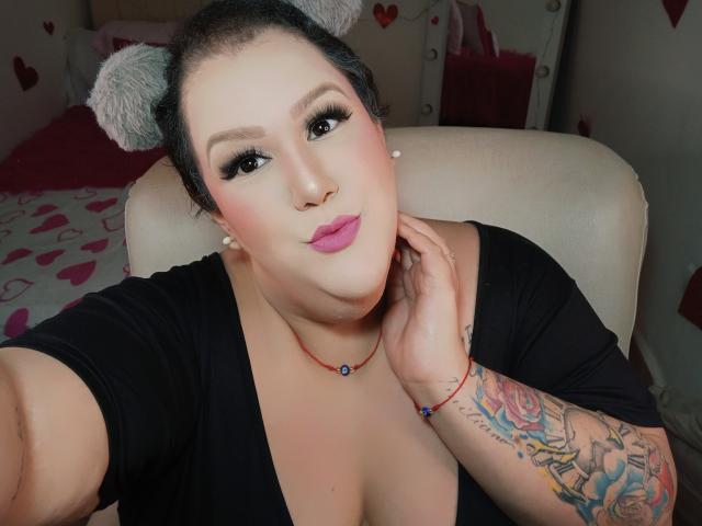 GiiannaMorrys, de piel clara y cabello oscuro, lleva una diadema gris de imitación de piel, una camiseta negra, lápiz labial rosa y tiene un tatuaje en el brazo. collar, mano en el cuello, expresión seductora, dormitorio tenuemente iluminado, patrones de corazones..