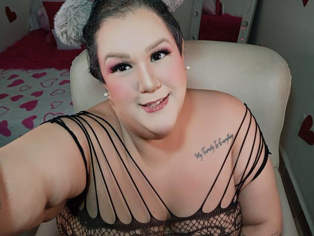 GiiannaMorrys, curvy and plump, with fair skin and black hair in a bun, wearing a revealing black mesh top, showing cleavage and a "Forever Young" tattoo on her chest. Smiling, pink eyeshadow.