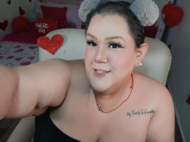 GiiannaMorrys, curvy woman with fair skin, black hair in gray bunny ears, red necklace, black strapless top, tattoo on left shoulder, smiling, red heart sticker, bedroom with heart decorations.
