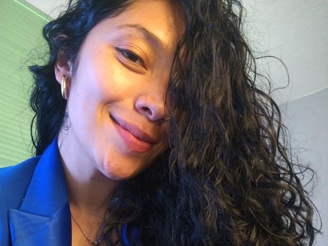 Close-up of ChantalBernadette, a smiling woman with long, curly black hair partially covering her face, wearing a blue blazer and gold earrings. Warm light highlights her slightly flushed skin. Fucking attractive.