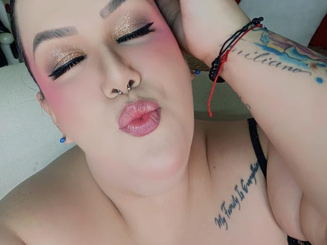 Close-up of GiiannaMorrys with heavy makeup, including pink blush and glittery gold eyeshadow. She has a nose ring, tattoos on her left arm and chest, and puckered lips. Black strap visible.