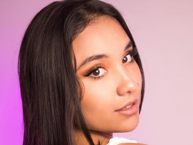 Close-up photo of BelleLane with long black hair, brown eyes, and a subtle smile. She's looking over her shoulder with a slight sexy gaze, pink and purple background. Fucking gorgeous.