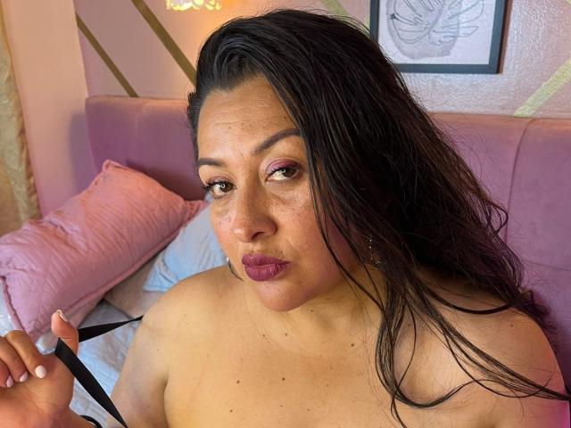 EvelynMatture with long black hair, dark brown eyes, and full lips, topless, holding a black strap, pink pillow in background, purple headboard, framed picture, sexy, confident expression, bedroom setting.