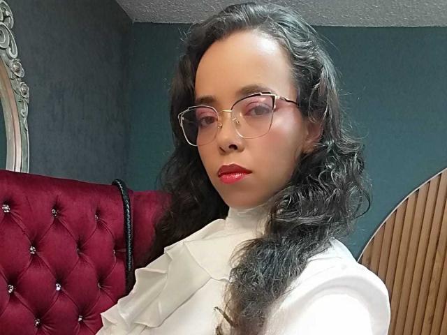 BarbaraLeythonn, a fair-skinned woman with long black wavy hair, wears glasses and a white shirt. She's seated on a red tufted chair, lips painted red, exuding confident, sexy allure.