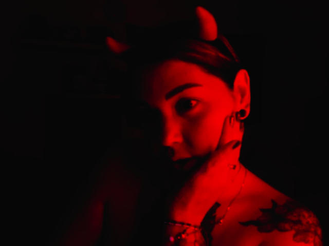 Photo en gros plan d'AlinaMatei, visage éclairé de rouge avec des ombres sombres, portant un serre-tête avec de petites cornes de diable, tatouage sur l'épaule. regard intense, reflets rouges sur la peau, fond noir, ambiance sensuelle et mystérieuse..