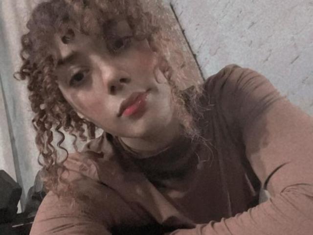 Primo piano di LioraVelvetC con capelli castani ricci, pelle chiara e labbra rosse, che indossa una camicia marrone a maniche lunghe e si appoggia. guardando in avanti, sguardo diretto, sottile tensione sessuale, bordi leggermente sfocati.