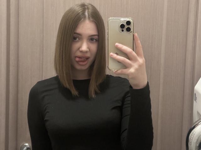 Elmorinaa, com cabelo castanho na altura dos ombros, a língua ligeiramente para fora, vestindo uma blusa preta justa de manga comprida, segurando um objeto dourado. iPhone, tirando uma selfie no espelho, fundo com painéis de madeira, com uma expressão sedutora..