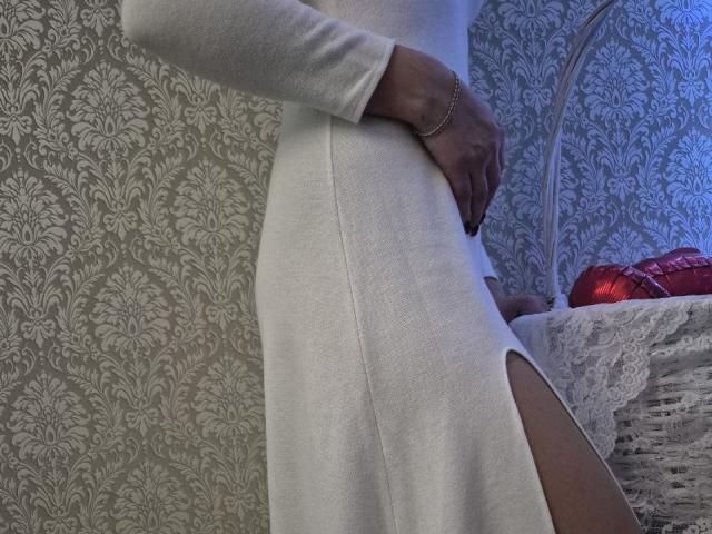 Anshante, a dark-skinned person, wears a tight white dress with a side slit, hand on belly, patterned wallpaper background, red glittery ornaments on lace-covered table, blue light on right.