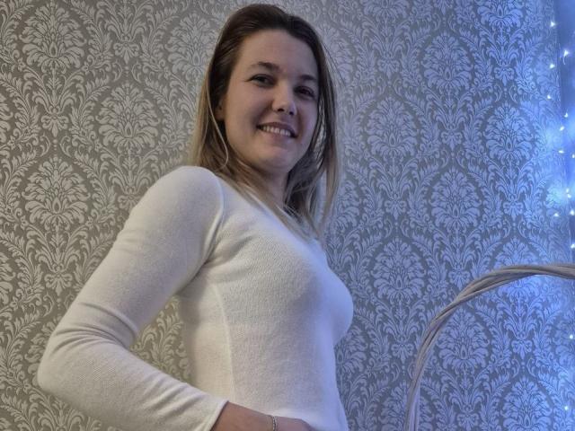 Anshante stands confidently in a white, long-sleeve top, smiling at the camera. She has light brown hair and a slender build. The background features ornate, silver-patterned wallpaper. Blue light illuminates the right side.