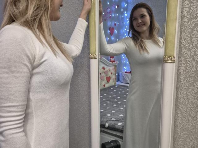 Anshante with blonde hair and a white, form-fitting dress stands in front of a mirror, smiling. She touches her hair with her left hand, reflecting in a mirror with gold accents. Blue and red heart lights in the background.