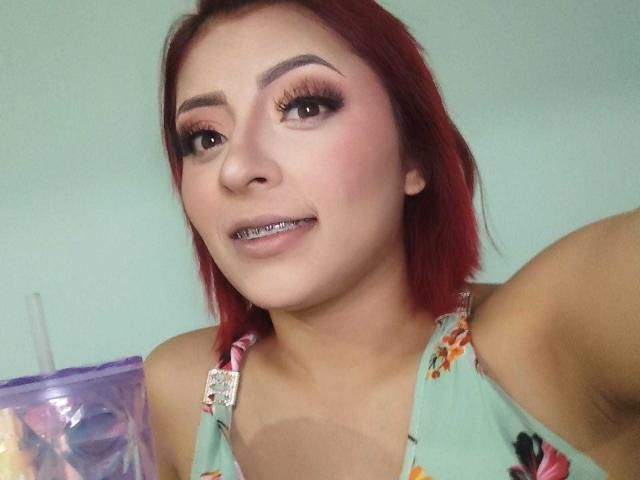 Selfie de primer plano de LizBonnet con cabello rojo, maquillaje intenso y delineador de ojos oscuro. Ella lleva una camiseta sin mangas con estampado floral, que deja entrever un poco su escote. Su expresión es ligeramente seductora..