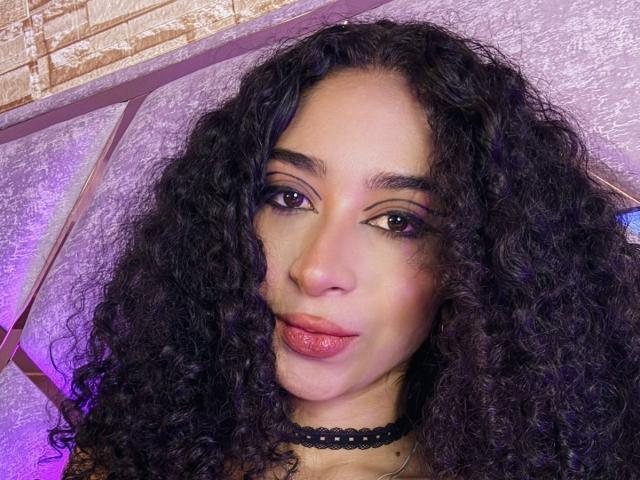 Close-up photo of LannaJhonson with curly black hair, dark eyes, and pink lips. She wears a black choker, with purple and pink lighting behind her. Fucking sexy look, direct gaze.