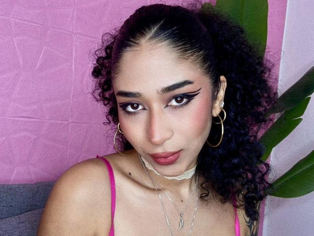 Foto em close-up de Lanna Jhonson, uma mulher curvilínea com cabelos cacheados escuros, delineador preto marcante, batom rosa e brincos de argola. brincos. Ela veste uma blusa rosa, um colar e olha sedutoramente para a câmera..